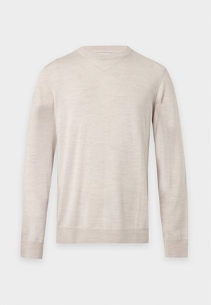 Jeune homme debout portant un pull beige clair, un pantalon marron et des chaussures noires sur un fond blanc uni.