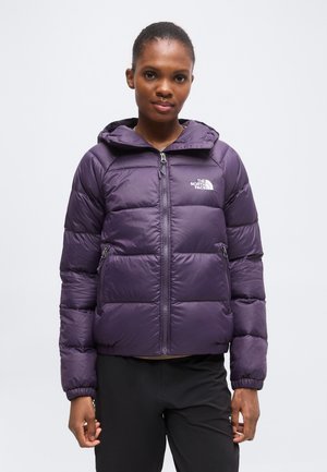 Donna che indossa una giacca imbottita viola The North Face con cappuccio, in piedi davanti a uno sfondo grigio chiaro uniforme.