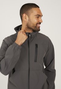 Grijze lichtgewicht jas met kap, rits aan de voorkant, elastische manchetten en een borstzak met een zwarte rits. Gladde, matte textuur.