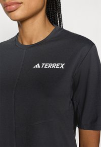 Čierne funkčné tričko s krátkym rukávom a okrúhlym výstrihom, s bielym logom "TERREX" na ľavej hornej strane. Hladká tkanina s detailmi švov.