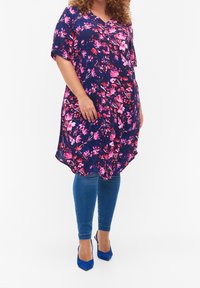 Robe tunique florale en marine avec des fleurs roses, manches courtes, décolleté en V et coupe ample, associée à un jean skinny bleu et des talons bleus.