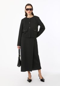 Veste noire courte avec poches avant et boutons métalliques, associée à une jupe assortie jusqu'aux chevilles. Accessoirisée avec des lunettes de soleil noires et des chaussures.