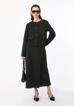 Black cropped jacket with front pockets and metallic buttons, paired with a matching ankle-length skirt. Styled with black sunglasses and shoes.