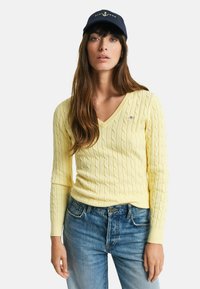 V-Ausschnitt, langärmliger Strickpullover in Pastellgelb mit strukturiertem Zopfmuster. Kombiniert mit blauen Jeans und einer marineblauen Mütze mit Logo.