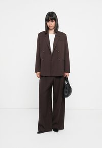 Brauner doppeltknöpfiger Blazer mit großen Revers, kombiniert mit weit geschnittenen Hosen. Darunter ein weißes Oberteil, hält eine kleine schwarze Handtasche.