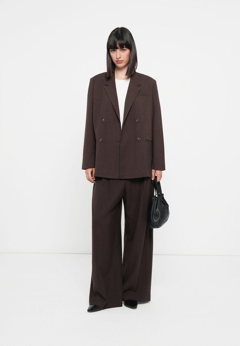 Brązowa dwurzędowa marynarka z dużymi klapami, w zestawie z szerokimi spodniami. Pod spodem biała bluzka, trzymająca małą czarną torebkę.