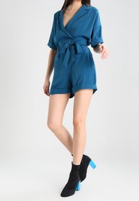 Teal omlottbyxa med korta ärmar och bälte, tillverkad av ett mjukt tyg. Kombineras med svarta ankelstövlar med klara blå blockklackar.