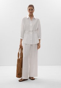 Chemise en lin blanche avec taille à cordon, accompagnée d'un pantalon ample assorti. Le modèle tient un sac tissé marron et porte des tongs marron.