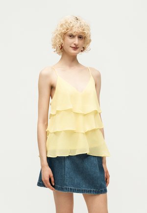 Femme aux cheveux blonds bouclés portant un haut jaune sans manches à volants et une mini-jupe en jean bleue, debout devant un fond uni.