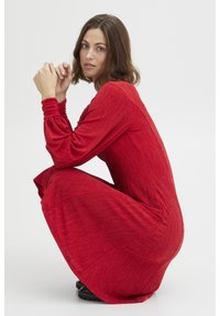 Röd klänning i mellanlängd med långa ärmar, texturerad tyg och elastiska manschetter. Modellen sitter på huk, vilket visar en avslappnad passform och sidofickor.