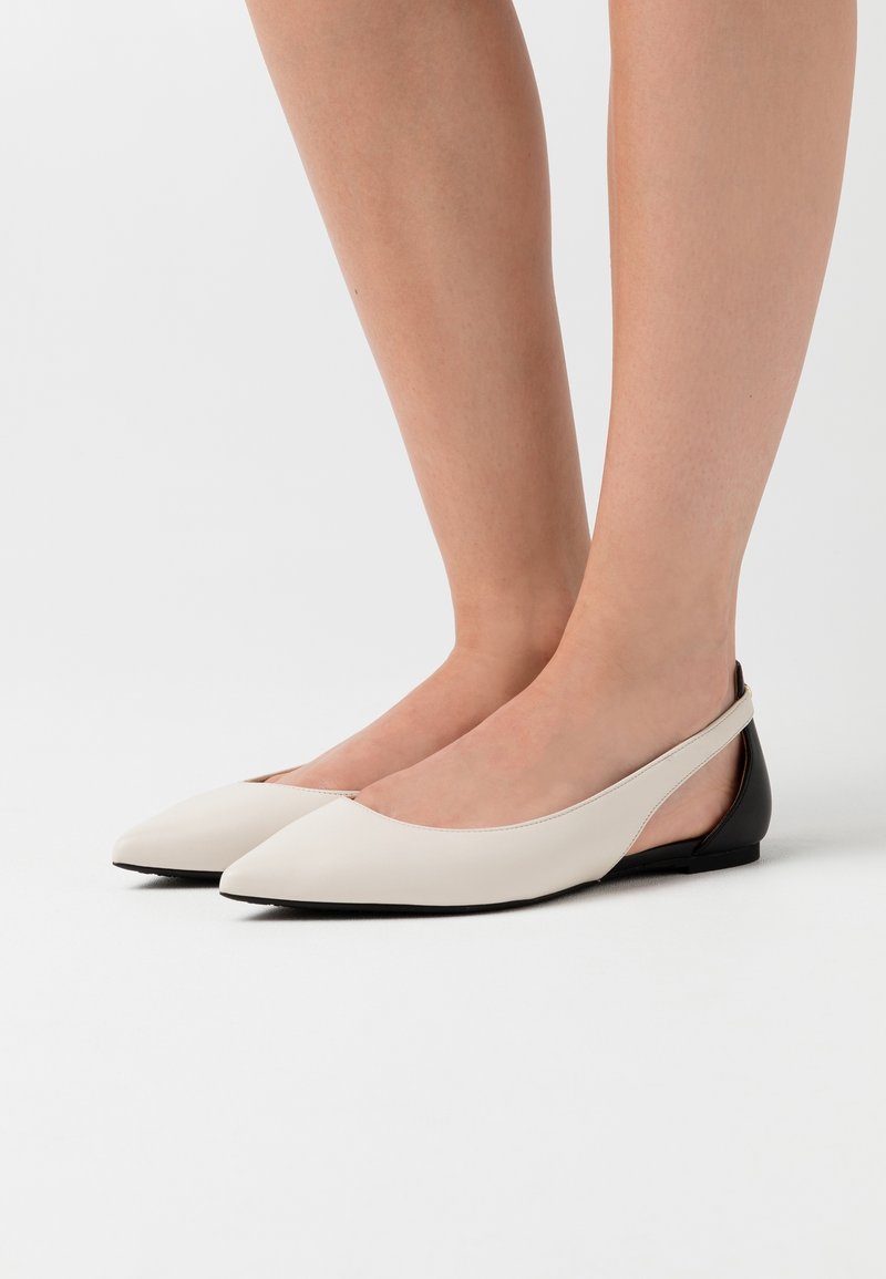 Des chaussures plates en cuir beige avec un bout pointu, un design à bride arrière et un accent de talon noir. Texture lisse, style minimaliste et profil bas.