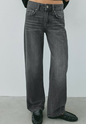 Kvinde iført sort, højtaljede wide-leg jeans med falmet effekt, kombineret med sorte slip-on sko, stående på en grå gulv mod en almindelig væg.