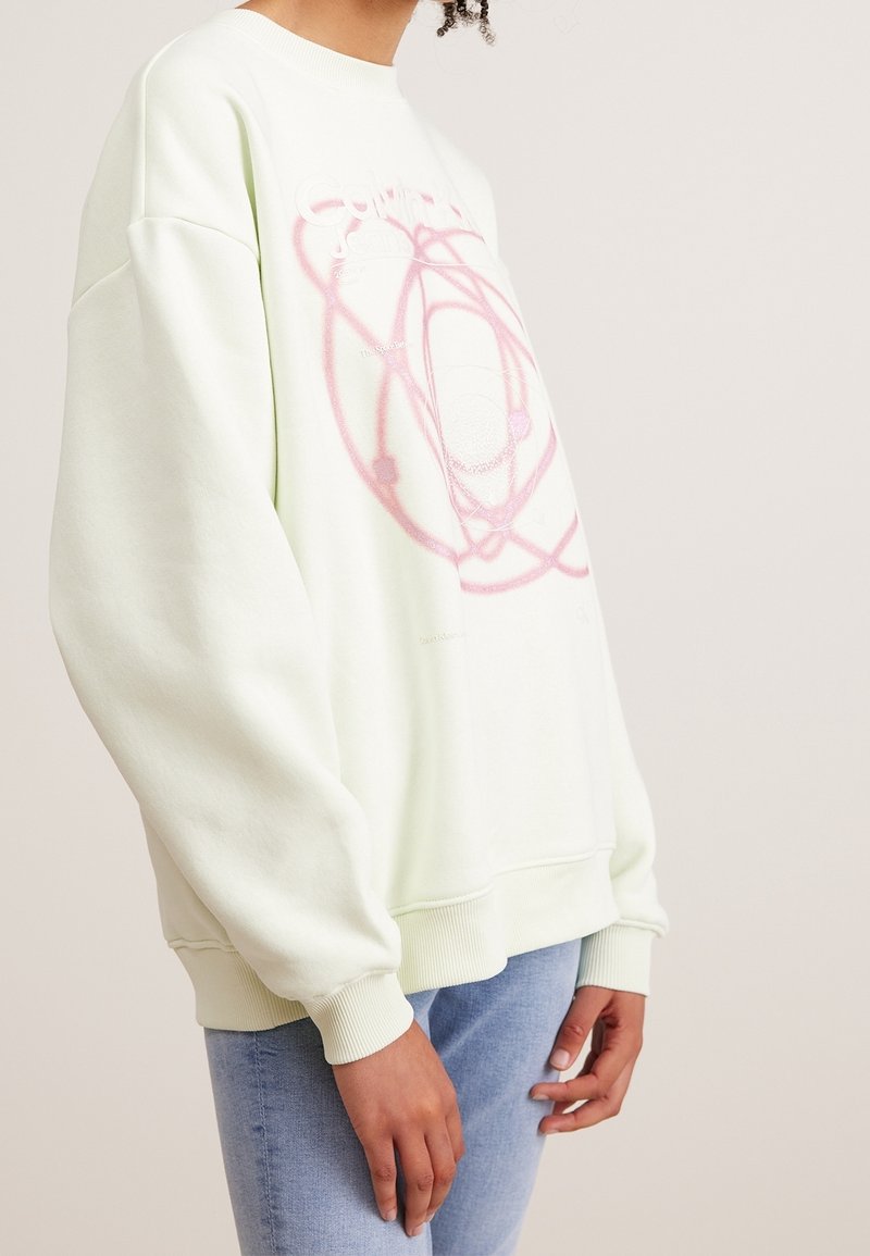 Personne portant un sweat-shirt oversize vert clair avec un motif abstrait rose et un jean bleu clair, debout devant un fond clair uni.