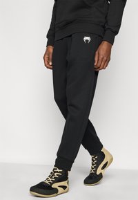 Μαύροι αθλητικοί παντελόνες με ριμπ μανσέτες, με λευκό λογότυπο στο μηρό. Συνδυάζονται με μπεζ και μαύρα αθλητικά παπούτσια ψηλής κορυφής.