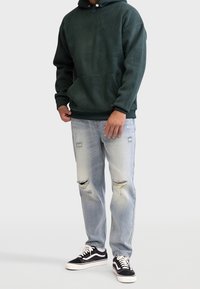 Mørkegrøn fleece hættejakke med en kengurulomme, kombineret med lyse, slidte denimjeans og sorte sneakers med hvide accenter.