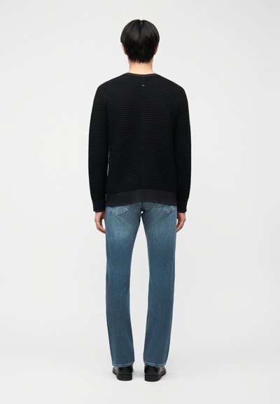 Άνδρας που στέκεται γυρισμένος με την πλάτη, φοράει μαύρο υφασμάτινο πουλόβερ με μακριά μανίκια, μπλε τζιν με ίσια μπατζάκια και μαύρα παπούτσια, σε λευκό φόντο.