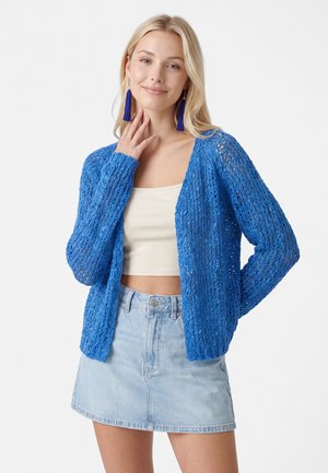 Ung kvinde iført en blå åbenstrikket cardigan, hvid crop top, lys denimsnørk og lange blå kvastøreringe, smilende med hånden nær ansigtet.
