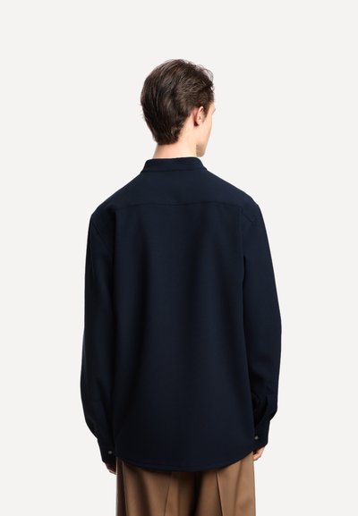 Uomo vestito con una camicia larga di colore blu navy scuro e pantaloni marroni, di spalle contro uno sfondo bianco semplice.