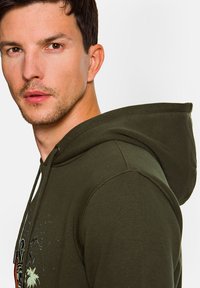 Oliwkowa bluza z kapturem z regulowanym sznurkiem, miękka w dotyku, z graficznym wzorem z przodu przedstawiającym palmy i ptaki. Luźny krój z dużym kapturem.