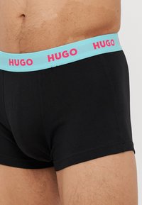 Schwarze Boxer-Shorts mit einem hellblauen elastischen Bund, der mit pinkem "HUGO"-Schriftzug versehen ist. Glatte Textur und figurbetontes Design.