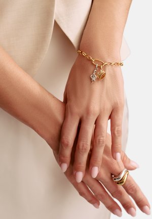 Goldene Gliederarmband mit Anhängern: silberne Schlange, goldener Ring und Krone. Glatte Oberfläche mit polierter Ausführung, getragen an einer Hand mit hellrosa lackierten Nägeln.