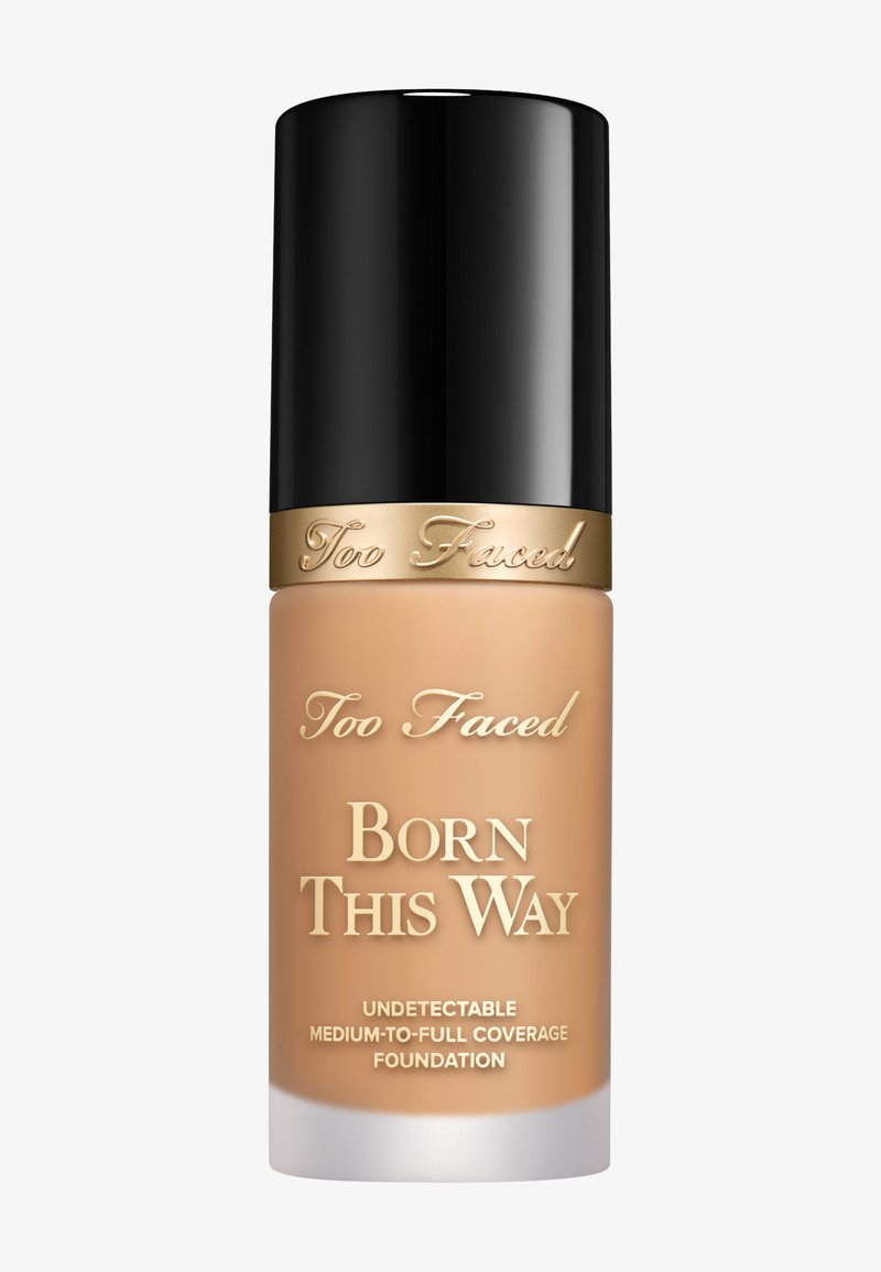 Too Faced Born This Way foundation in een doorzichtige glazen fles met een zwarte dop. Het etiket heeft gouden tekst op een beige achtergrond.