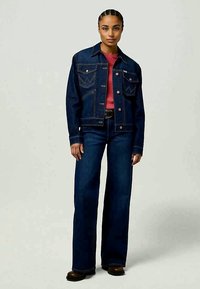 Casaco de ganga azul escuro com um corte clássico, apresentando bolsos frontais e costura visível. Combinado com jeans flare a condizer e uma camisa vermelha.