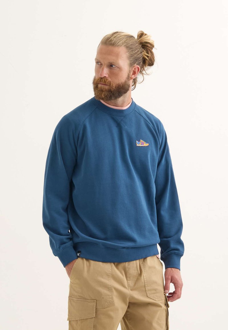 Felpa blu con colletto a coste e polsini a coste, con un piccolo grafico ricamato. Indossata con pantaloni cargo beige.
