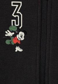 Abbigliamento nero con il numero "3" bianco e una grafica colorata di Topolino in rosso e verde. Il tessuto sembra liscio con un dettaglio a zip.