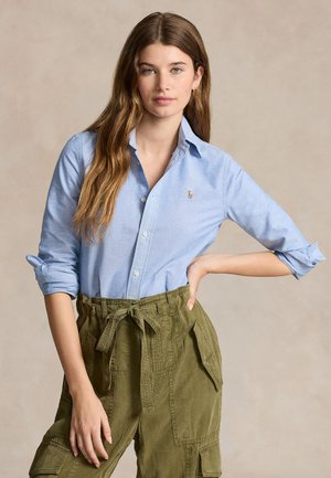 Ung kvinne med langt brunt hår iført en lyseblå skjorte med knapper og olivengrønne høytlivsbukser med knytt belte.