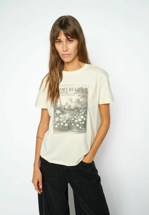 Vrouw met lang bruin haar draagt een crèmekleurig T-shirt met zwart-witte bloemen- en gebouwenprint en een zwarte broek, staand tegen een witte achtergrond.
