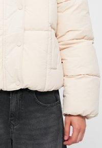 Kremfarget boblejakke med cropped design, som har store quiltede seksjoner og trykknapplukking. Brukt over mørke denimbukser.