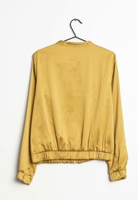 Gelber Satin-Bomberjacke mit elastischen Bündchen und Saum, die an einem schwarzen Kleiderbügel vor einer wei�ßen Wand hängt.