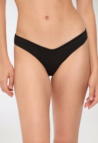 Tanga in cotone nero con taglio a vita bassa, caratterizzato da una texture liscia e un davanti a forma di V. Cuciture minime e nessun dettaglio hardware visibile.