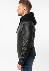 Schwarze Lederjacke mit Kapuze, taillierte Passform, seitliche Reißverschlusstaschen und strukturierte Schulterakzente. Getragen über einer blauen Jeansjacke mit strukturierter Oberfläche.