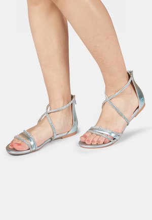 Srebrne metalliczne sandały z wieloma cienkimi paskami, płaską podeszwą i zdobione cyrkoniami. Posiadają zamek błyskawiczny z tyłu.