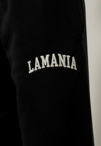 Juodas audinys su baltais siuvinėtais žodžiais "LAMANIA" didžiosiomis raidėmis, išlenktais per paviršių.