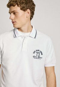 Balta polo krekla ar tumši zilu akcentu, divu pogu atloku un izšūtu logotipu "Pepe Jeans London 73" uz krūtīm. Īsas piedurknes.