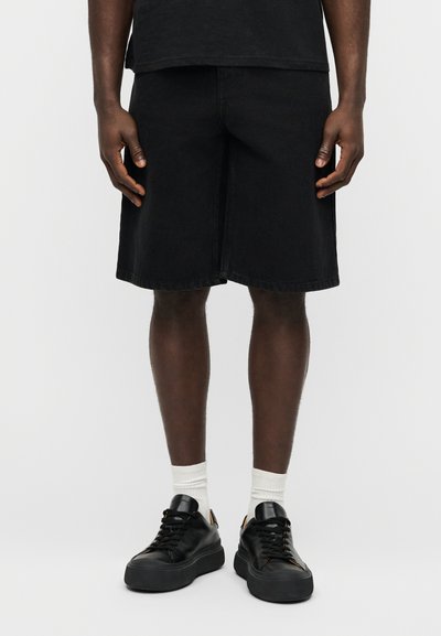 Μαύρα σορτς από τζιν με χαλαρή εφαρμογή, ευθεία γραμμή, ξεφτισμένο τελείωμα και διακριτικές λεπτομέρειες ραφής, φορεμένα με μαύρα αθλητικά παπούτσια και λευκές κάλτσες.