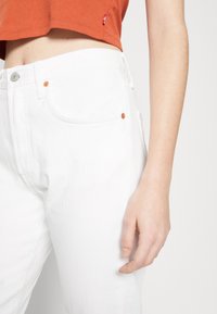 Jeans de algodón blancos con un diseño de pierna recta, que presentan detalles en botones de color cobre y costuras en contraste. Textura suave visible.
