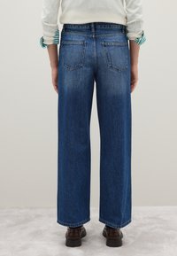 Weite blaue Denim-Jeans mit Fünf-Taschen-Design, distressed zum Saum hin und sichtbaren Nähten. Die Schuhe sind im braunen Lederlook.