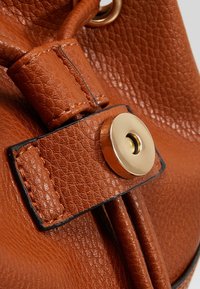 Borsa in pelle marrone con texture, dotata di chiusura a bottone automatico dorato e dettagli cuciti, presenta una parte superiore con cordoncino e una fodera interna liscia.