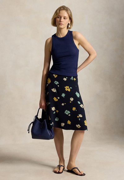 Jeune femme posant, portant un haut sans manches bleu marine, une jupe midi noire à fleurs, des sandales noires, tenant un sac à main bleu marine sur un fond neutre.