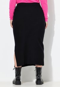 Een persoon draagt een zwarte midi-rok met een split aan de achterkant, zwarte stevige laarzen en een langemouwige roze top met golvende randdetails.