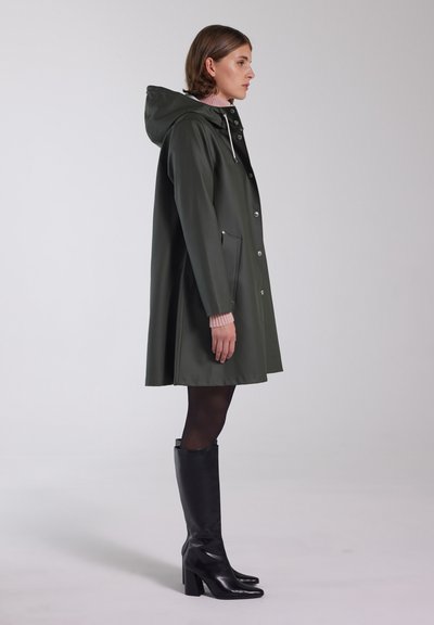 Groene waterdichte jas met een capuchon, zilveren drukknoopsluiting, zijzakken en een relaxte pasvorm. Draagbaar met kniehoge zwarte laarzen en zwarte panty.