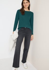 Langarm-Türkisoberteil kombiniert mit dunkelgrauen weiten Jeans und weißen Sneakern. Das Modell hält in einer Hand einen hellgrauen Pullover.