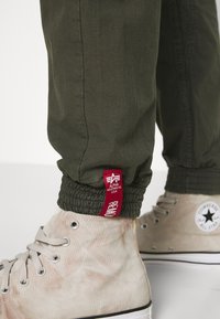 Olivové joggery s textúrovanou látkou, elastickými manžetami a červenou značkovou etiketu. Na nohách sú svetlé, béžové vysoké tenisky.