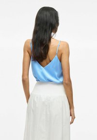 Modrá saténová košieľka s nastaviteľnými ramienkami a tvarom V na chrbte, v kombinácii s bielou sukňou so smockovaným pásom. Hladká textúra, jednoduchý dizajn.
