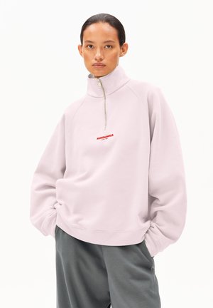 Junge Person trägt oversized Pullover in Hellrosa mit halbem Reißverschluss und rotem "ARMEDANGELS"-Logo, dunkelgraue Hose, steht mit Händen in den Taschen.