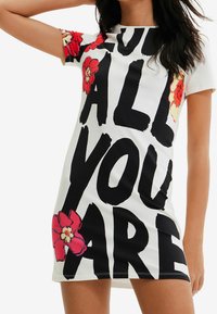 Šaty s krátkým rukávem z bílé bavlny s výrazným černým textem "MILUJ VŠECHNO, CO JSI" a červenými květinovými akcenty na rukávech a dolním lemu.